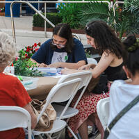 Several people sit at outdoor tables, drawing or crafting together, surrounded by plants and flowers.