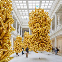 Large clusters of gold balloons float in a spacious, white museum hall with high ceilings and people walking around.
