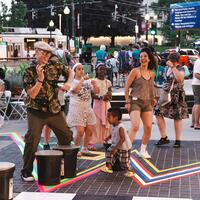 People of various ages dance and play drums outdoors at a lively community event on a city street.