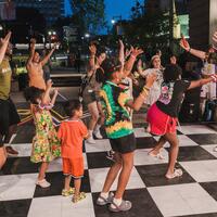 A group of adults and children dance energetically together outdoors on a black and white checkered floor at night.