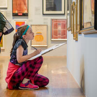 A young girl kneels on the floor sketching art in a museum gallery, focused on framed artworks on the wall.