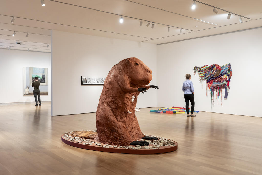 A beaver-shaped sculpture in the center with two people in the background looking at artwork in a museum.