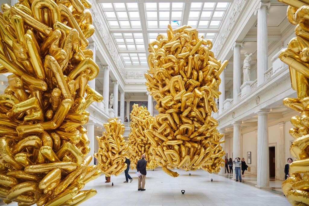 Large clusters of gold balloons float in a spacious, white museum hall with high ceilings and people walking around.