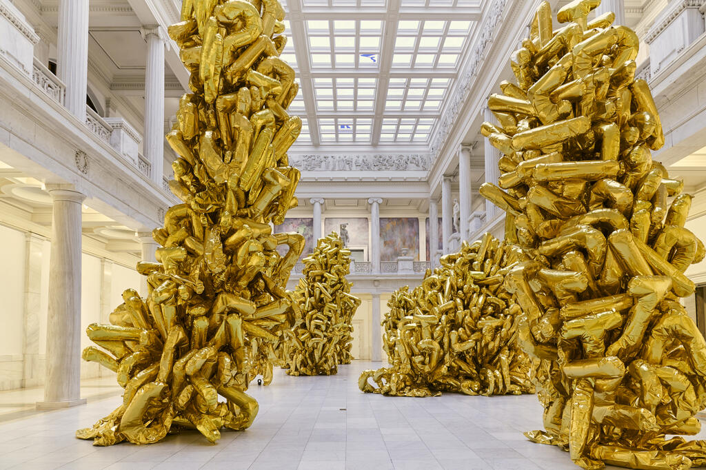 Tall clusters of shiny gold inflated sculptures fill a grand, white marble hall with a glass ceiling.