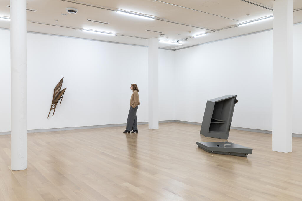 A person stands in a gallery between a wall-mounted wooden sculpture and a large geometric floor installation.