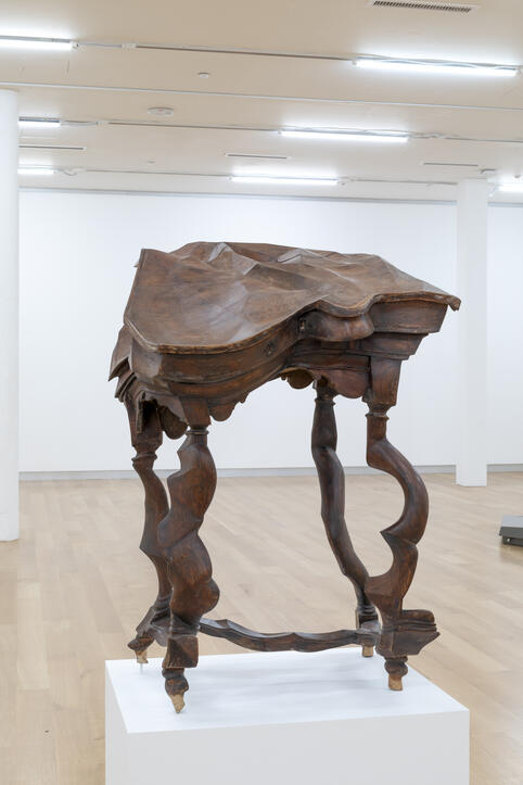 A sculptural, warped wooden table with distorted, twisted legs displayed on a white pedestal in a gallery.