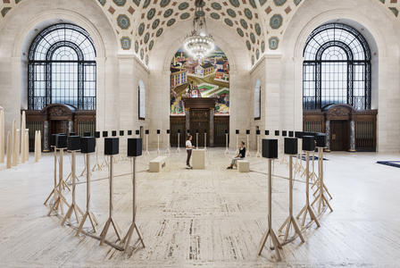 An installation view of 40 speakers on plain wooden stands arranged in a circle with two visitors listening