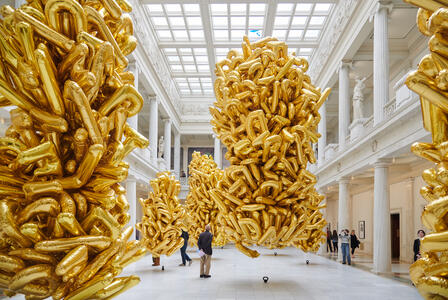 Large clusters of gold balloons float in a spacious, white museum hall with high ceilings and people walking around.