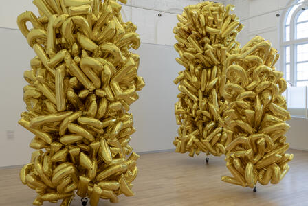 Three tall clusters of shiny gold balloons stand in a bright, modern gallery with wooden floors.