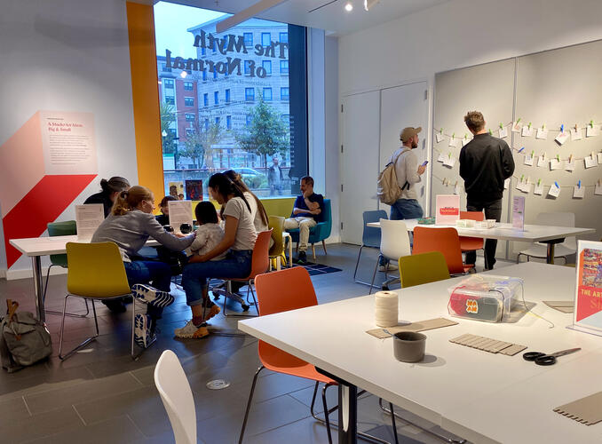 A group of people sitting and standing in a room with arts and crafts tools on a white table.