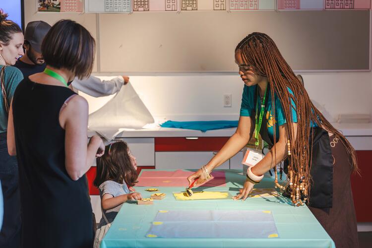 Student showing printmaking technique to a young child with adults standing nearby.