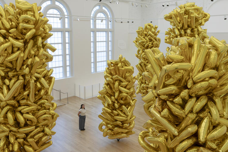 A person stands among tall clusters of gold balloon sculptures in a bright, spacious gallery.