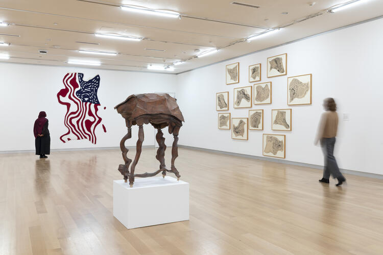 Visitors walk through an art gallery featuring a wooden sculpture on a pedestal, a flag mural, and framed drawings.