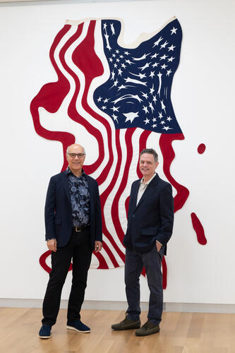 Two men in blazers standing in front of a large, abstract felt textile of the American flag.