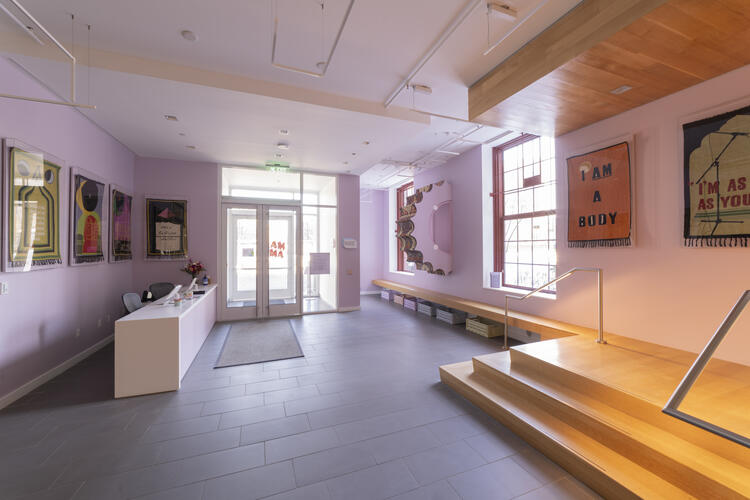 Interior of a purple-walled gallery featuring a reception desk, textile wall hangings, and a wooden staircase.