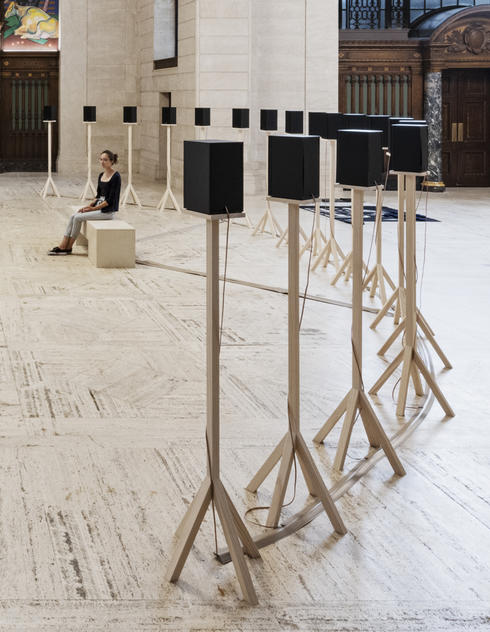 Installation view of speakers on plain wooden stands and a seated visitor listening