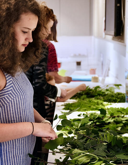 A young woman arranges green leaves on a table in a bright room with others working beside her.