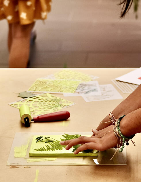 A person arranges leaves on a printing plate at a craft table with art supplies and paper nearby.