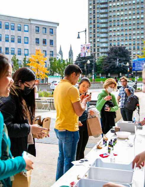 People stand in line at an outdoor table with supplies, smiling and talking, with city buildings in the background.