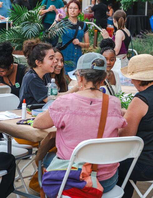 A group of people sit around a table outdoors, talking and crafting at a community event.