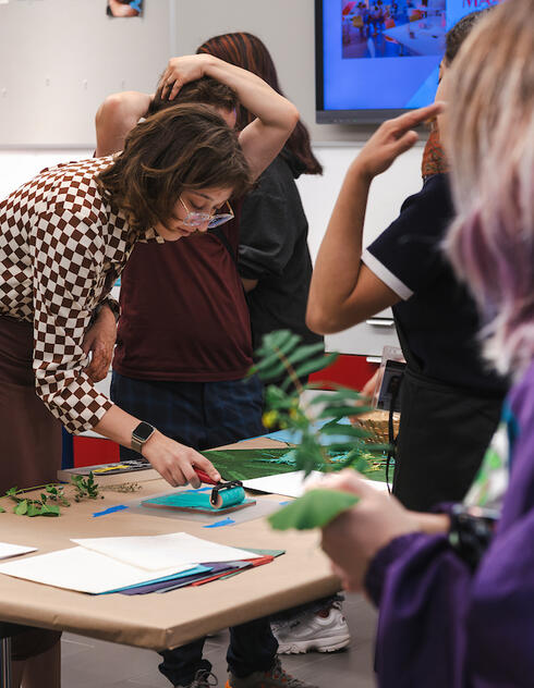 A woman demonstrates art techniques at a table while several people observe and participate in a classroom setting.