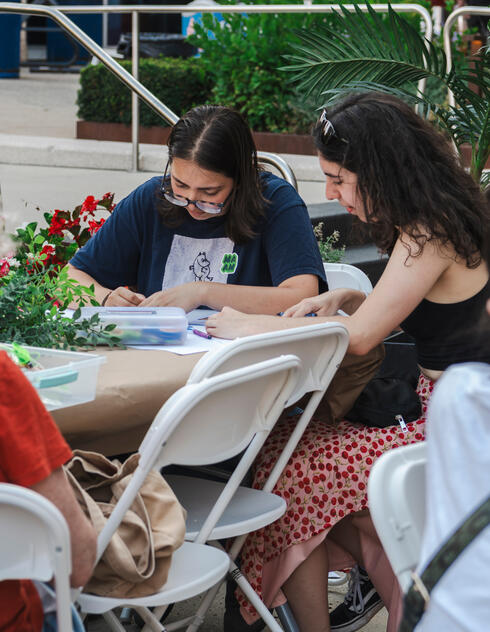 Several people sit at outdoor tables, drawing or crafting together, surrounded by plants and flowers.