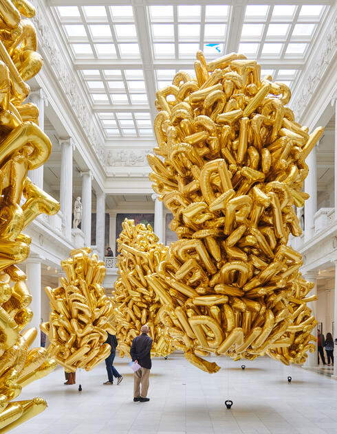 Large clusters of gold balloons float in a spacious, white museum hall with high ceilings and people walking around.