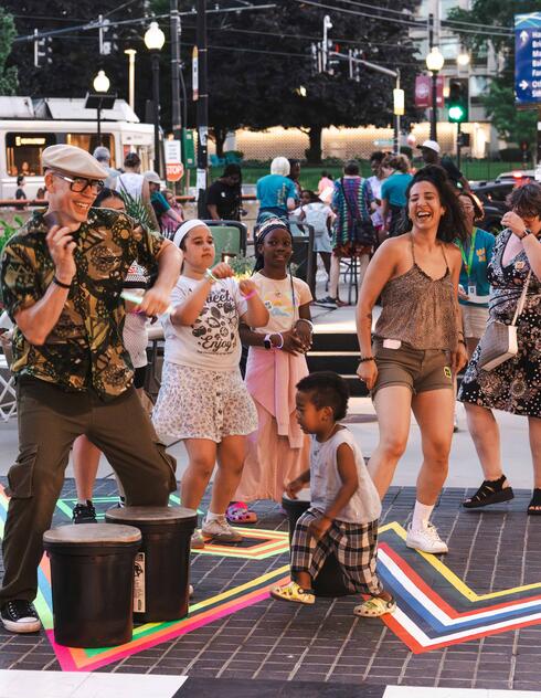 People of various ages dance and play drums outdoors at a lively community event on a city street.