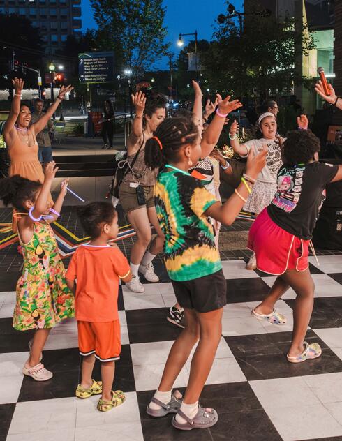 A group of adults and children dance energetically together outdoors on a black and white checkered floor at night.