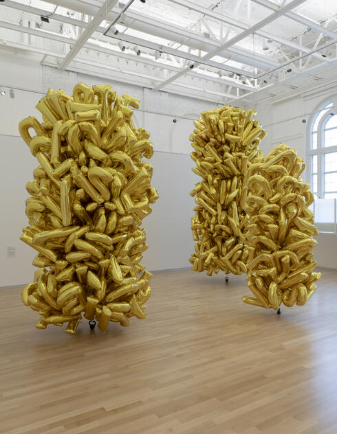 Three tall clusters of shiny gold balloons stand in a bright, modern gallery with wooden floors.