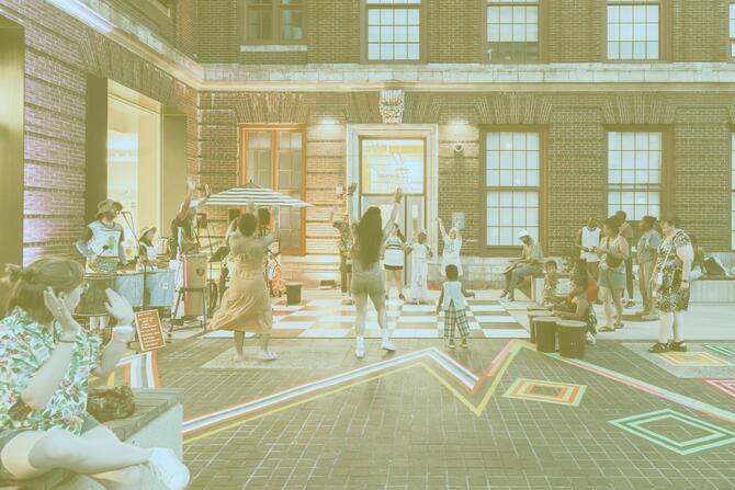 People dance and watch a street performance on a brick plaza in front of a building with large windows.