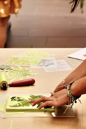 A person wearing colorful bracelets arranges fern leaves on paper for a printmaking project at a table.
