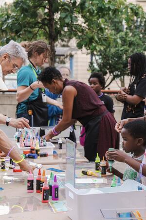 Children and adults gather around a table outdoors, doing arts and crafts with colorful paint bottles and supplies.