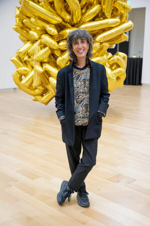 A person in a black blazer and patterned shirt stands in front of a large, gold, abstract sculpture.
