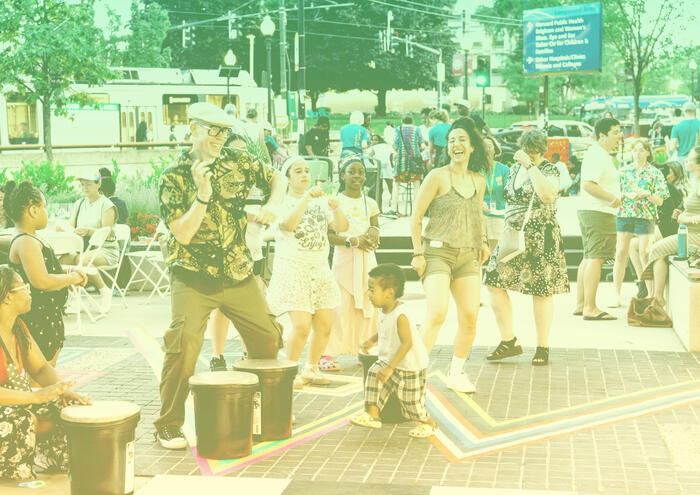 People of all ages dance and play drums outdoors at a lively community event on a city street.