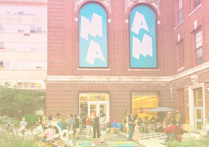 People gather outside a brick building with large blue MAN banners and white chairs arranged on the sidewalk.