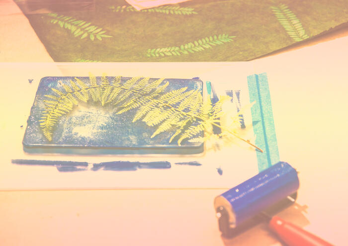 A fern leaf on an inked plate with a roller, fabric, and leaf prints in the background.