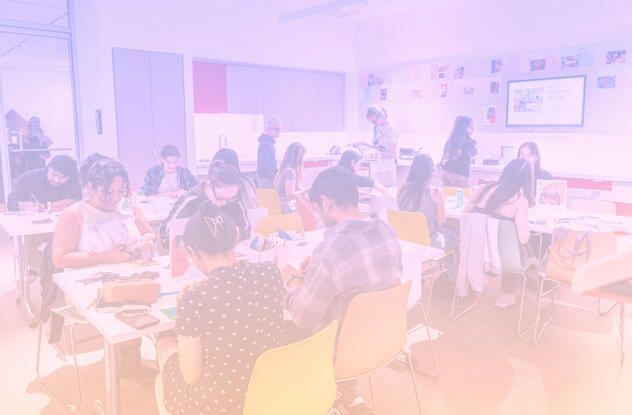 A brightly lit workspace with people around tables, engaged in art activities. Walls are adorned with colorful artwork, creating a lively, creative atmosphere.